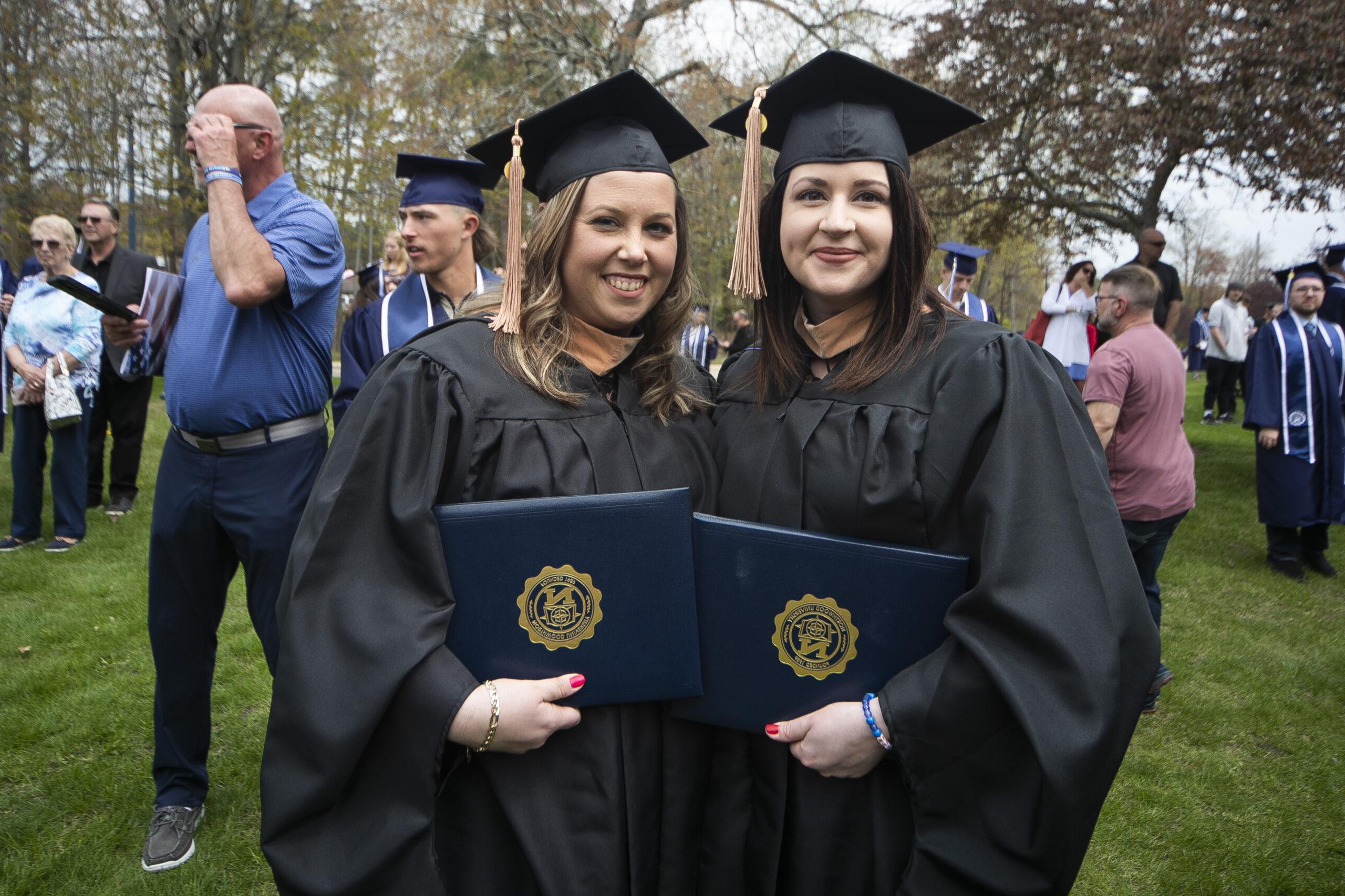 两名竞技宝app下载大学的毕业生戴着学士帽，在外面举着他们的毕业证书